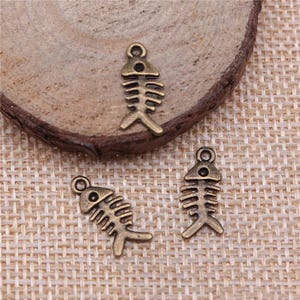 May include: Three antique bronze-toned metal charms shaped like fish skeletons. Each charm features a small circular loop at the top for attachment, a triangular head section with a single hole, and a series of horizontal ribs ending in a tail.