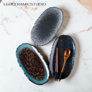 Pode incluir: Três pratos ovais de cerâmica com desenhos salpicados em azul escuro e branco. Um prato tem uma borda turquesa. Pauzinhos e uma colher de pau repousam sobre um prato. O texto "LEOCERAMICSTUDIO" está no topo.