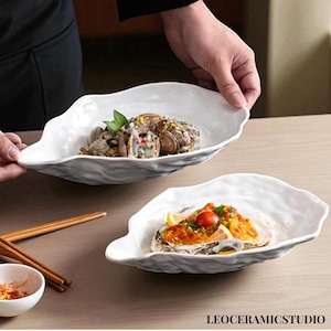 May include: Two white, organically shaped ceramic serving dishes. One dish holds cooked clams with herbs, the other holds a seafood dish with a tomato garnish. The dishes are on a light brown surface. The text "LEOCERAMICSTUDIO" is visible.