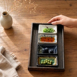 May include: A wooden serving tray with four small rectangular ceramic dishes. The dishes contain food items like seaweed, sauce, blueberries, and peas. The tray is on a wooden table, and a hand is holding the tray.