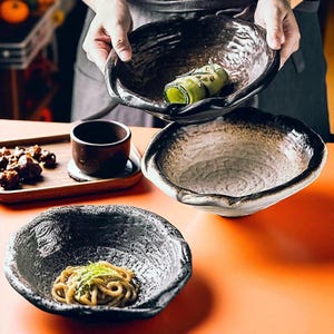 May include: Handmade ceramic bowls with a rustic, textured design. The bowls are dark with a lighter interior and irregular edges. One bowl holds noodles, another a cucumber roll, and a third is empty. A small brown cup and wooden tray are also visible.