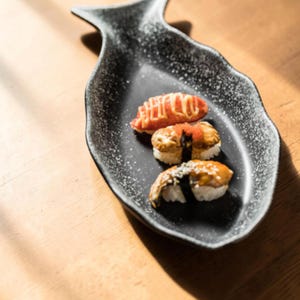 Puede incluir: Un plato de cerámica negro con forma de pez con tres piezas de sushi. El sushi incluye una pieza con un aderezo rojo, una con un aderezo marrón y otra con un aderezo oscuro. El plato tiene un diseño moteado.