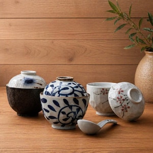 Op de afbeelding: Een verzameling Japanse keramische kommen en een lepel. De kommen hebben ingewikkelde blauw-witte patronen, met een donkerbruine basis voor één kom. Eén kom heeft een deksel. De lepel is wit.