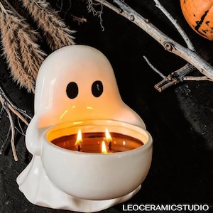 Könnte beinhalten: Ein weißer Keramik-Geist-Kerzenhalter mit zwei schwarzen Augenausschnitten. Der Halter enthält eine Dreidochtkerze mit warmem Schein. Der Text "LEOCERAMICSTUDIO" ist unten sichtbar. Ein Dekorationsartikel zum Thema Halloween.