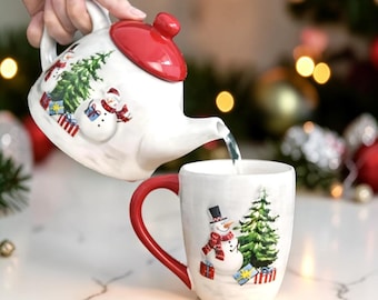 May include: A white ceramic teapot with a red lid is pouring liquid into a matching mug. Both the teapot and mug are decorated with a snowman, Christmas tree, and gift boxes. The mug has a red handle. The text "LEOCERAMICSTUDIO" is at the bottom.