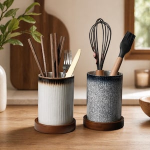 Może przedstawiać: Dwa ceramiczne pojemniki na przybory na drewnianych podstawach. Jeden jest biały z brązowym wykończeniem i zawiera pałeczki, widelec i nóż. Drugi jest niebiesko-szary z trzepaczką i pędzlem.