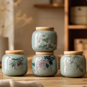 Puede incluir: Cuatro pequeños tarros redondos de cerámica con tapas de madera están dispuestos sobre una superficie de madera. Los tarros son de color verde claro con diseños florales en blanco, rosa y rojo. Los tarros están apilados y dispuestos para crear una exhibición decorativa.