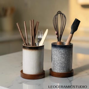 Op de afbeelding: Twee cilindrische keramische keukengerei-houders op ronde houten voetjes. De linkerhouder is wit met verticale ribbels en een donkere rand, gevuld met houten eetstokjes, een metalen vork en een mes. De rechterhouder is grijs gespikkeld met een donkere rand, gevuld met een garde en een siliconen bakkwast. De tekst luidt LEOCERAMICSTUDIO.