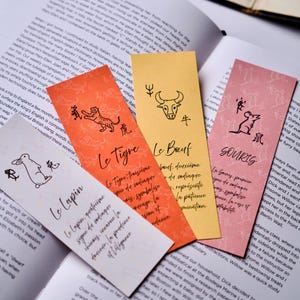 Może przedstawiać: Cztery kolorowe zakładki do książek z ilustracjami zwierząt zodiaku i tekstem w języku francuskim. Zakładki są pomarańczowe, żółte, różowe i białe i leżą na otwartej książce. Każda zakładka ma inne zwierzę.