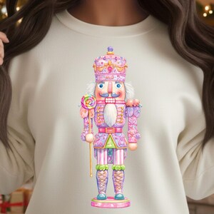 May include: Cream-colored sweatshirt featuring a colorful nutcracker design. The nutcracker is depicted in pink, purple, and blue with a crown and a lollipop. The sweatshirt is part of a holiday-themed collection.