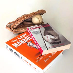 May include: A baseball glove, baseball, and two books are displayed on a white surface. The books are titled "The Last Manager" and "Moneyball" by Michael Lewis. The baseball is white with red stitching.