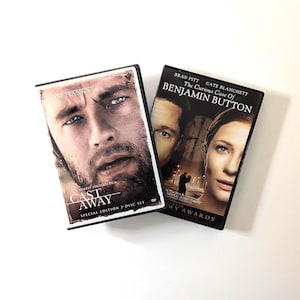 May include: Two rectangular DVD cases featuring movie cover art. The left case displays the title Cast Away with a close-up of a bearded man. The right case shows The Curious Case of Benjamin Button with portraits of a man and a woman.