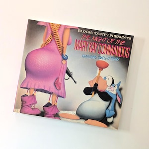 May include: A square Bloom County print titled "The Night of the Mary Kay Commandos" featuring cartoon characters. The artwork depicts a figure in a pink dress with a machine gun and a cartoon penguin holding a plunger. The text "FEATURING SMELL-O-TOONS" is also visible.