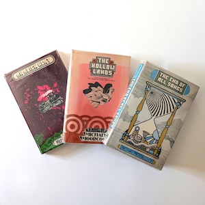 May include: Three vintage paperback books with colorful covers. Titles include "An Alien Heat," "The Hollow Lands," and "The End of All Songs." The covers feature illustrations of a train, a bird, and an hourglass.