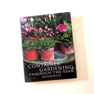 May include: A hardcover book titled Container Gardening Through the Year by Malcolm Hillier. The cover features a photograph of three terracotta pots containing various green plants and pink flowers, set on a weathered circular stone surface.