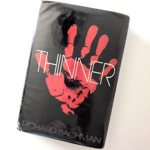 May include: A paperback book with a black cover featuring a large red handprint and the title "THINNER" in white. The author's name, Richard Bachman, is printed at the bottom. The book is a novel.