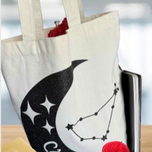 May include: White canvas tote bag with black Capricorn zodiac design. The bag contains a red ball of yarn, a black notebook, and a small yellow wallet. The bag has two cream-colored handles.