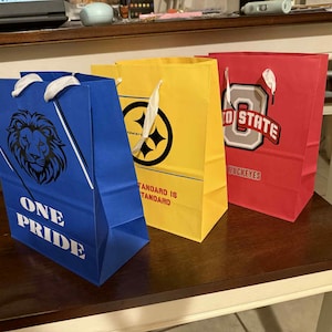 Könnte beinhalten: Drei farbige Geschenktüten: blau, gelb und rot. Die blaue Tasche hat eine Löwengrafik und die Worte "ONE PRIDE". Die gelbe Tasche hat einen schwarzen Kreis mit einem Sterndesign. Die rote Tasche hat ein weißes "O" mit "STATE" darüber und "BUCKEYES" darunter.