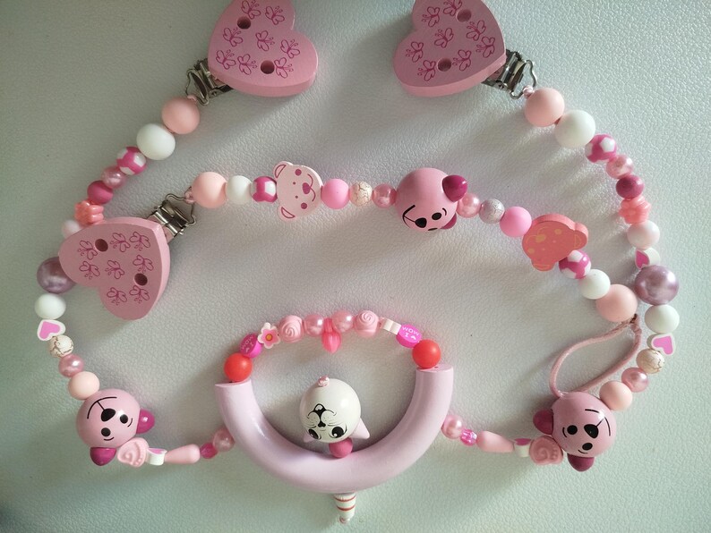 May include: Baby pacifier clips in shades of pink, featuring heart and bear motifs. The clips are adorned with pink, white, and pearl beads, and include a curved teething ring with a white animal figure.
