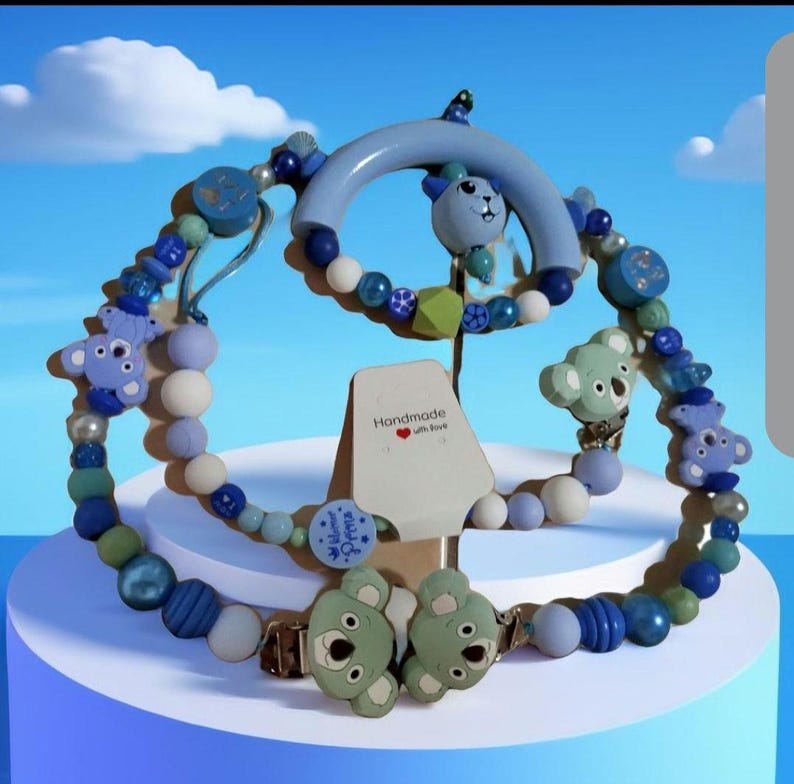 K&ouml;nnte beinhalten: Ein Schnullerclip- und Bei&szlig;ring-Set f&uuml;r Babys in Blau-, Gr&uuml;n- und Wei&szlig;t&ouml;nen. Das Set zeigt Koala-B&auml;ren- und Perlen-Designs mit dem Schriftzug "Handmade with love" auf einem Etikett. Der Schnullerclip hat einen Metallverschluss.