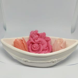 Könnte beinhalten: Eine weiße Keramikschale enthält drei dekorative Seifen. Eine rosa Seife in Form eines Engels liegt auf einer Rose, flankiert von zwei tulpenförmigen Seifen in Rosa und Pfirsich. Die Schale hat eine Bootsform.