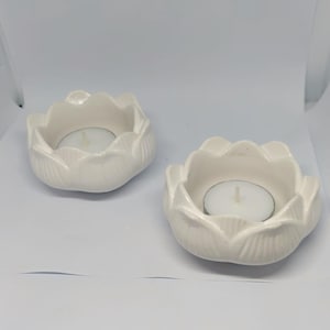 Könnte beinhalten: Zwei weiße Keramik-Lotusblüten-Kerzenhalter, jeweils mit einer kleinen weißen Teelichtkerze. Die Halter haben ein Blütenblatt-Design. Der Hintergrund ist weiß.