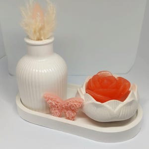 Könnte beinhalten: Ein weißes Keramiktablett enthält eine kleine weiße Vase mit flauschigen beigefarbenen Federn, einen rosa Schmetterling und eine weiße Keramikschale in Rosenform mit einer roten Rose darin. Die Artikel sind auf einer weißen Oberfläche angeordnet.