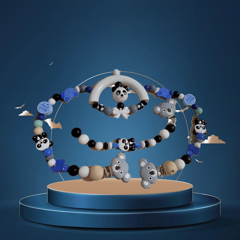 K&ouml;nnte beinhalten: Ein Babymobile mit einem wei&szlig;en Bogen, einem Panda und Schn&uuml;ren aus blauen, schwarzen, wei&szlig;en und grauen Perlen. Das Mobile enth&auml;lt Panda- und Koala-B&auml;renfiguren sowie kleine blaue Perlen in Form von Autos. Das Mobile wird auf einer blauen Plattform pr&auml;sentiert.