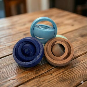 May include: Three wooden, spherical toys in shades of blue and brown are arranged on a wooden surface. One toy has a handle, while the others have concentric circles. The toys are designed with a unique, geometric aesthetic.
