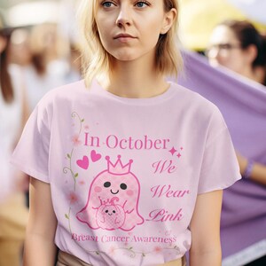 Op de afbeelding: Lichtroze T-shirt met de tekst "In October We Wear Pink" en een afbeelding van een roze spook met een kroon en een pompoen. Het shirt is voor borstkankerbewustzijn.