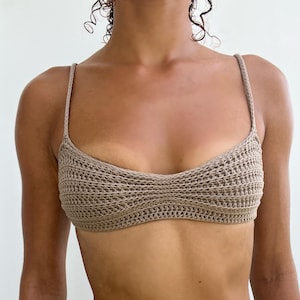 Peut inclure: Haut en crochet beige avec fines bretelles. Le haut a un aspect texturé et tissé et une encolure dégagée. L'image montre le haut sur une personne, avec un tatouage sur le bras gauche et un piercing au nombril.