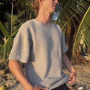 Può includere: Una maglietta a maniche corte con girocollo realizzata in un tessuto a maglia all'uncinetto color beige chiaro testurizzato. Il capo presenta un motivo a punti coerente e ripetitivo su tutto il corpo e le maniche, offrendo un aspetto strutturato.