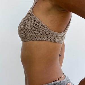 May include: A tan crochet top with a halter neck design. The top is made of a textured, open-weave material. The image shows the side view of the top, highlighting the intricate crochet pattern and the overall shape. The top is a neutral color.