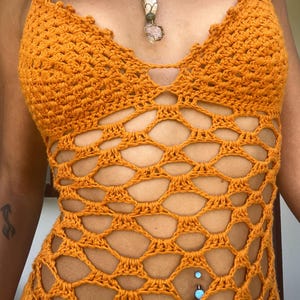 May include: A crochet halter top in a vibrant orange hue. The top features a unique openwork design with a scalloped neckline and a halter strap. A necklace with various gemstones hangs from the neck. The top is a handmade garment.