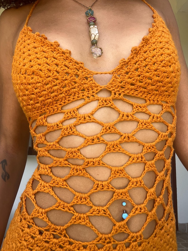 May include: A crochet halter top in a vibrant orange hue. The top features a unique openwork design with a scalloped neckline and a halter strap. A necklace with various gemstones hangs from the neck. The top is a handmade garment.