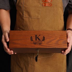 May include: A dark brown wooden box with a key lock, held by a person wearing a brown apron. The box is engraved with the letter "K", laurel leaves, and the name "Kristian". The wood has a rich, dark brown finish.