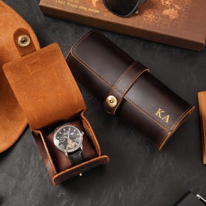 Könnte beinhalten: Ein braunes Leder-Uhrenetui mit einer Uhr darin, neben einem gerollten Leder-Uhrenetui mit den Initialen "KA" in Gold. Sonnenbrillen und eine Schachtel mit Weltkartenmuster sind ebenfalls zu sehen.