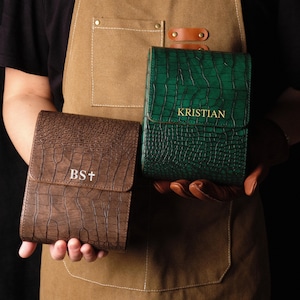 May include: Two leather cases with a crocodile skin texture. One is brown with "BS+" embossed, the other is green with "KRISTIAN" embossed. Both have a flap closure. The cases are held by a person wearing a brown apron.