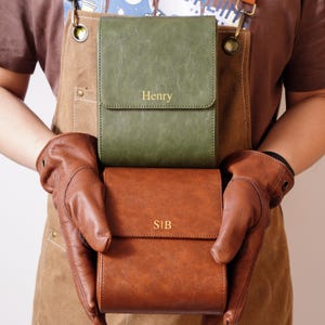 Puede incluir: Dos carteras de cuero, una verde oliva y otra marrón, se muestran. La verde tiene grabado "Henry", la marrón "SIB". Ambas tienen cierre de solapa. Las carteras son sostenidas por alguien con guantes de cuero marrón.