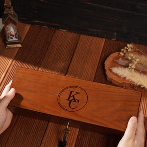 Peut inclure: Une bo&icirc;te en bois rectangulaire avec une serrure et une cl&eacute; avec un pompon noir. La bo&icirc;te pr&eacute;sente un embl&egrave;me circulaire avec les lettres "KC" grav&eacute;es. Une montre-bracelet dor&eacute;e est visible.