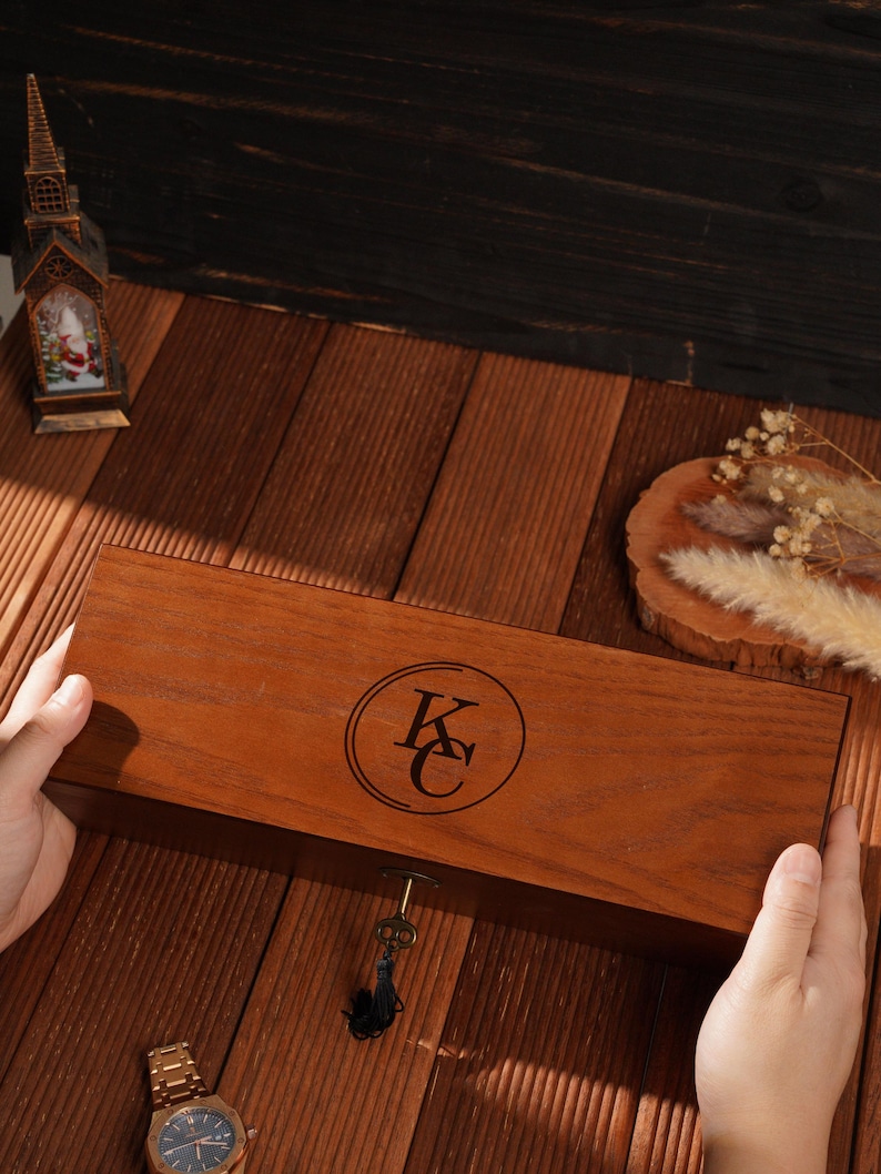 Peut inclure: Une bo&icirc;te en bois rectangulaire avec une serrure et une cl&eacute; avec un pompon noir. La bo&icirc;te pr&eacute;sente un embl&egrave;me circulaire avec les lettres "KC" grav&eacute;es. Une montre-bracelet dor&eacute;e est visible.