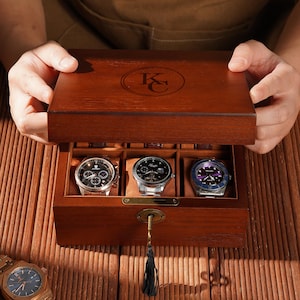 May include: A wooden watch box with three watches inside. The box is open, revealing the watches with different colored faces and bands. The box has a key and a tassel. A gold watch is on the wooden surface.