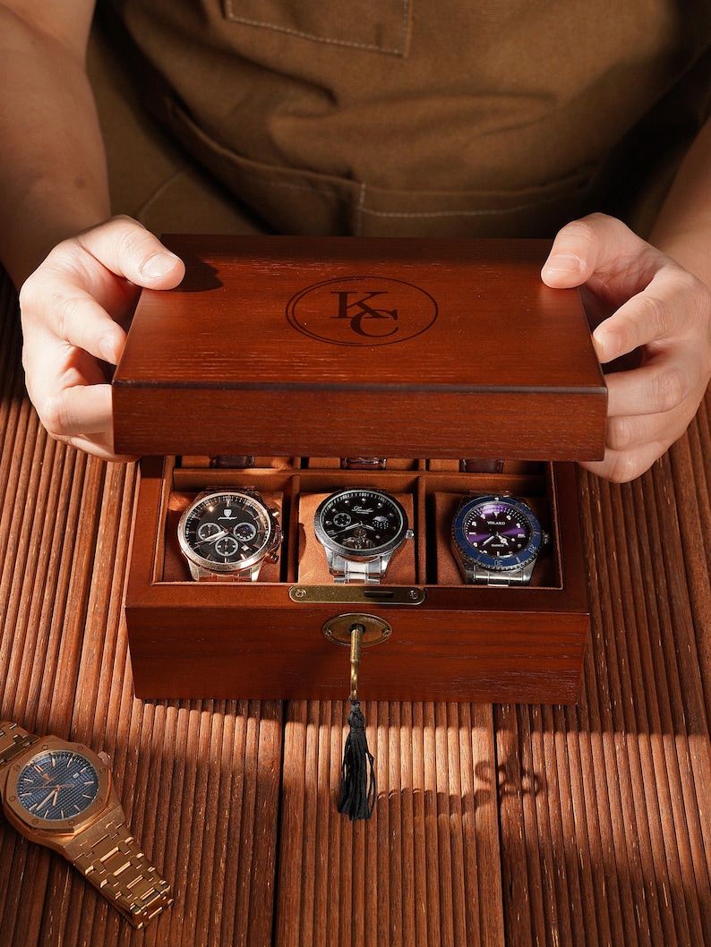 Peut inclure: Coffret en bois fonc&eacute; ouvert, r&eacute;v&eacute;lant trois montres avec diff&eacute;rents cadrans et bracelets. Le coffret est orn&eacute; d'une cl&eacute; et d'un pompon. Une montre dor&eacute;e est pos&eacute;e sur la surface en bois. Le couvercle porte un monogramme.