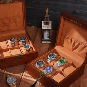 Peut inclure: Deux coffrets &agrave; montres en bois ouverts avec plusieurs montres &agrave; l'int&eacute;rieur. Les coffrets sont bruns avec un int&eacute;rieur beige. Une montre dor&eacute;e avec un cadran bleu est pos&eacute;e &agrave; l'ext&eacute;rieur d'un coffret. Une petite figurine d'&eacute;glise se trouve entre les coffrets.