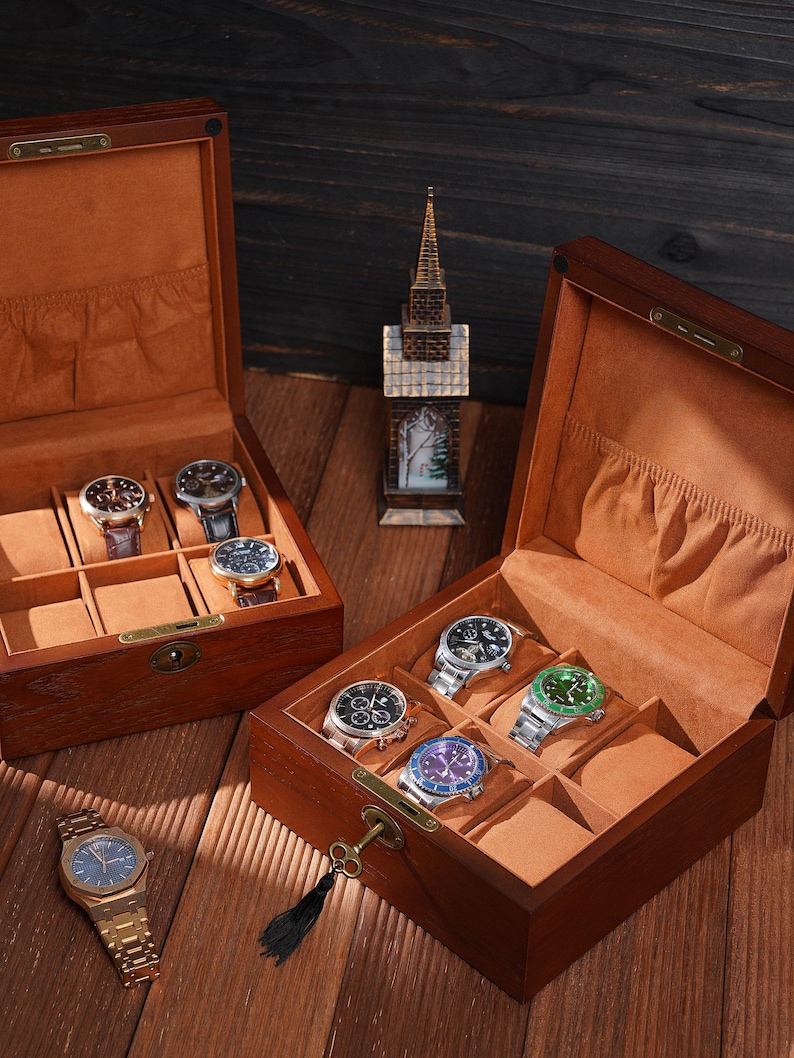 Peut inclure: Deux coffrets &agrave; montres en bois ouverts avec plusieurs montres &agrave; l'int&eacute;rieur. Les coffrets sont bruns avec un int&eacute;rieur beige. Une montre dor&eacute;e avec un cadran bleu est pos&eacute;e &agrave; l'ext&eacute;rieur d'un coffret. Une petite figurine d'&eacute;glise se trouve entre les coffrets.