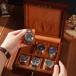 Peut inclure: Une bo&icirc;te &agrave; montres en bois avec un int&eacute;rieur marron, contenant plusieurs montres-bracelets avec diff&eacute;rents mod&egrave;les de bracelets et de cadrans. Une montre est tenue par une main. La bo&icirc;te est ouverte, r&eacute;v&eacute;lant les montres &agrave; l'int&eacute;rieur.