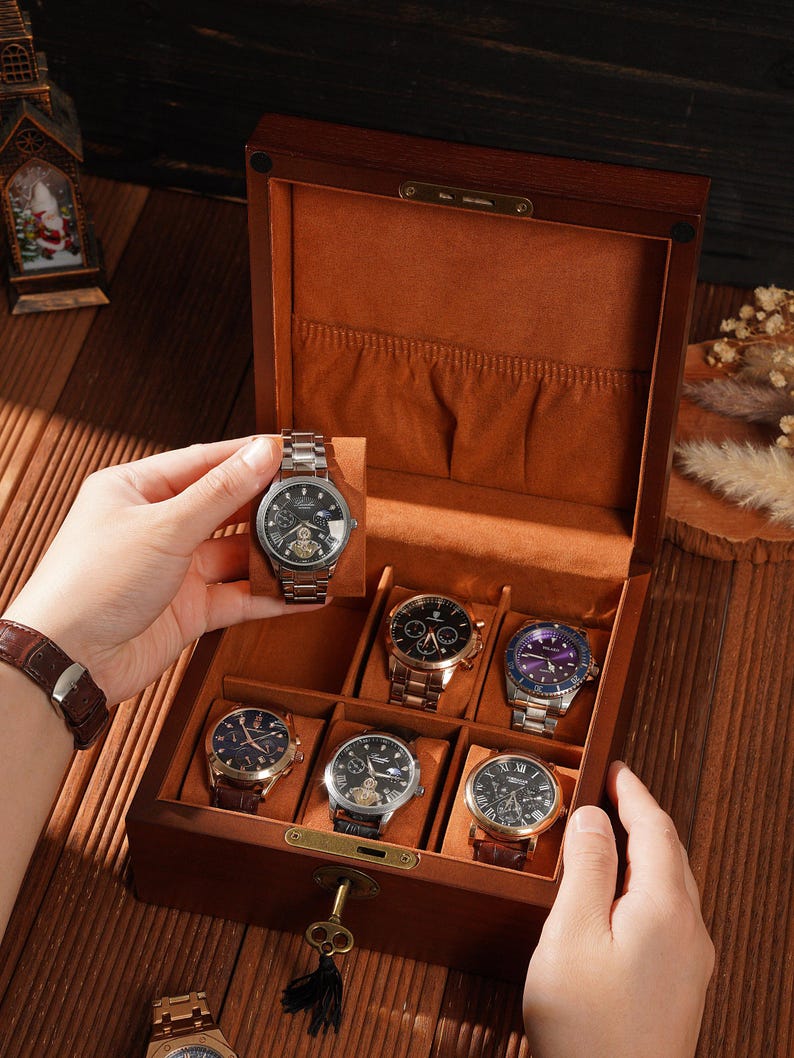 Peut inclure: Une bo&icirc;te &agrave; montres en bois avec un int&eacute;rieur marron, contenant plusieurs montres-bracelets avec diff&eacute;rents mod&egrave;les de bracelets et de cadrans. Une montre est tenue par une main. La bo&icirc;te est ouverte, r&eacute;v&eacute;lant les montres &agrave; l'int&eacute;rieur.