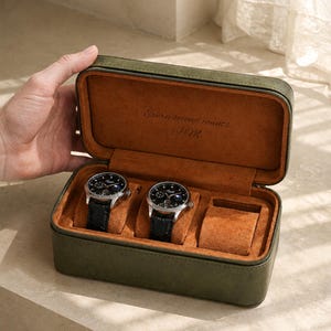 Puede incluir: Un estuche rectangular de color verde oliva para guardar relojes con un forro interior de color naranja suave. La tapa presenta el texto grabado "Every second counts J.M." y la base contiene dos relojes de pulsera plateados con correas negras.