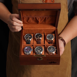 Peut inclure: Une bo&icirc;te en bois pour montres avec six compartiments, chacun contenant une montre. La bo&icirc;te est doubl&eacute;e de tissu orange et poss&egrave;de une serrure. Les montres ont diff&eacute;rentes couleurs de bracelets et de cadrans. La bo&icirc;te est personnalis&eacute;e avec le nom "KRISTIAN" et une moustache.