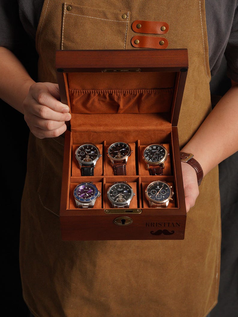 Peut inclure: Une bo&icirc;te en bois pour montres avec six compartiments, chacun contenant une montre. La bo&icirc;te est doubl&eacute;e de tissu orange et poss&egrave;de une serrure. Les montres ont diff&eacute;rentes couleurs de bracelets et de cadrans. La bo&icirc;te est personnalis&eacute;e avec le nom "KRISTIAN" et une moustache.